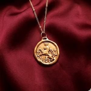 May include: A gold-toned pendant necklace featuring a circular coin-style charm with an embossed lion standing on its hind legs, topped with a small crown and surrounded by leafy branches, suspended from a delicate gold-toned metal chain.