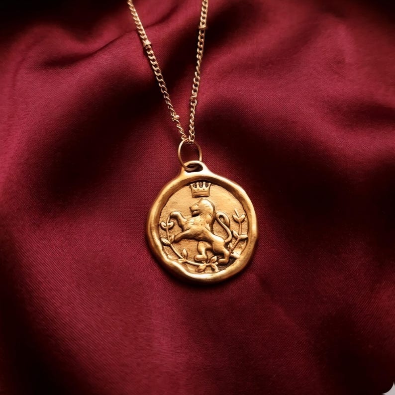 May include: A gold-toned pendant necklace featuring a circular coin-style charm with an embossed lion standing on its hind legs, topped with a small crown and surrounded by leafy branches, suspended from a delicate gold-toned metal chain.