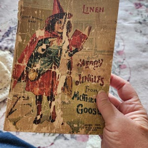 May include: A vintage 1902 linen book titled Merry Jingles From Mother Goose, part of the Little Miss Muffet Series. The cover features a color illustration of a child in a red hat and cape reading a book, with significant surface wear and paper loss.