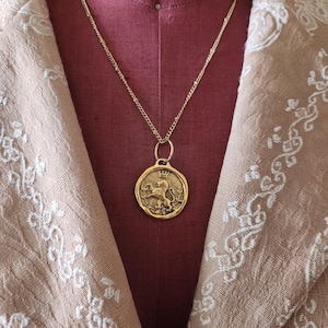 May include: A gold-toned necklace featuring a circular pendant with a raised relief design of a crowned lion standing on its hind legs, surrounded by a thin border, suspended from a delicate gold-toned chain with small, evenly spaced links.