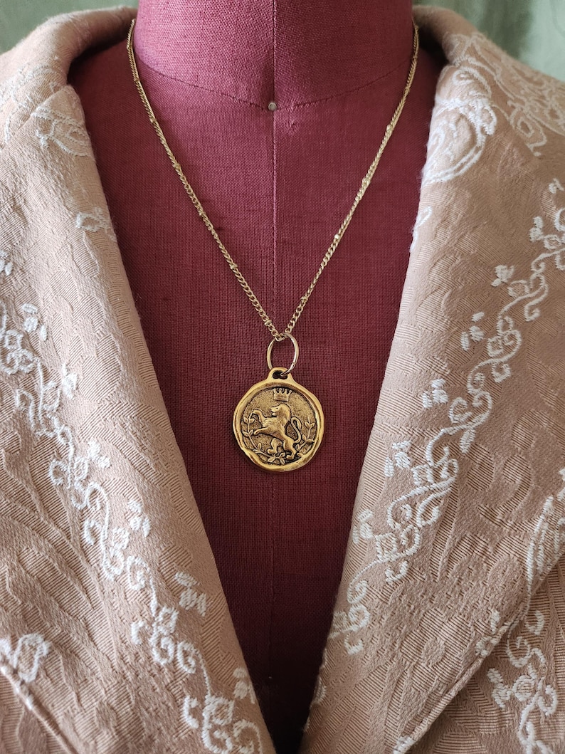 May include: A gold-toned necklace featuring a circular pendant with a raised relief design of a crowned lion standing on its hind legs, surrounded by a thin border, suspended from a delicate gold-toned chain with small, evenly spaced links.