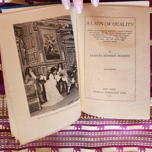 May include: An open hardcover book titled A Lady of Quality by Frances Hodgson Burnett, published in 1914 by Charles Scribner's Sons. The left page features a black and white illustration of figures in period clothing, while the right page displays text.