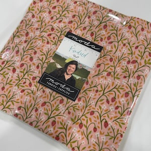 May include: A square stack of fabric squares featuring a pink background with a repeating pattern of green vines, small white flowers, and red strawberries. A label on top reads Moda Kindred, ICANDE2, and Moda Fabrics + Supplies, with a 4-inch ruler.