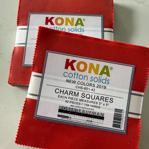 May include: Two stacks of red fabric squares labeled Kona Cotton Solids Charm Squares. Each pack contains 42 pieces measuring 5 inches by 5 inches, totaling .756 yards. The packaging includes the Robert Kaufman Fabrics logo and a barcode.