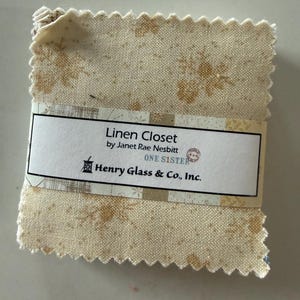 May include: A square stack of fabric squares with pinked edges, featuring a cream and tan floral pattern. A white paper band wraps around the center, printed with the text: Linen Closet by Janet Rae Nesbitt, ONE SISTER, Henry Glass & Co., Inc.