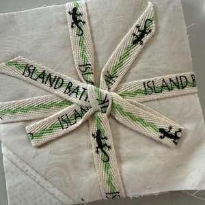 May include: A square stack of off-white fabric tied with a cream-colored ribbon featuring a repeating green zigzag pattern and black lizard silhouettes. The ribbon is printed with the text ISLAND BATIK in black capital letters and tied in a center knot.