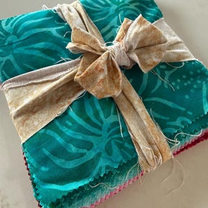 May include: A square stack of fabric squares with pinked edges, featuring a teal and green botanical leaf pattern. The stack is tied together with a tan, mottled-print fabric ribbon tied in a bow at the top center of the fabric bundle.