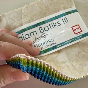 May include: A stack of 42 fabric squares labeled Alam Batiks III Pistachio by Jinny Beyer for RJR Fabrics. The fabric squares feature pinked edges and a gradient color palette ranging from cream and light green to vibrant green, teal, and deep blue.