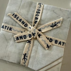 May include: A stack of square fabric pieces in light gray and off-white tones, tied together with a cream-colored ribbon. The ribbon features the repeating black text ISLAND BATIK AND BLK and small black lizard icons printed along its length.