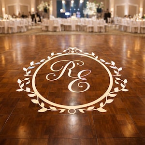 May include: A wooden dance floor with a large, circular monogram in the center. The monogram features the letters "R" and "E" in an elegant script, surrounded by a decorative wreath. The floor reflects the lights of a ballroom.