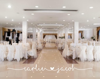 May include: A large, bright banquet hall featuring rows of tables covered in white tablecloths and chairs adorned with white fabric covers. The polished floor reflects the overhead lighting. The text 'carlie & jacob' is centered.