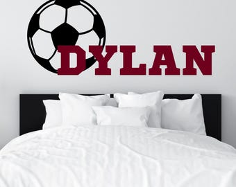 May include: A wall decal featuring a black and white soccer ball graphic positioned to the left of the name DYLAN, which is written in large, bold, maroon-colored capital letters in a collegiate-style block font.