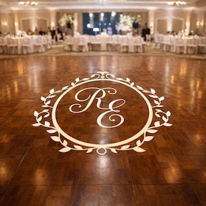 May include: A white monogram projection on a polished wooden dance floor featuring the letters RE in a cursive script, enclosed within a circular frame decorated with stylized leaves and vines, centered on the floor surface.