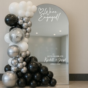 May include: An arched mirror with the text "We're Engaged!" and "Let's Celebrate!" with a couple's names and date. A balloon garland in black, silver, white, and a disco ball decorates the mirror's left side.