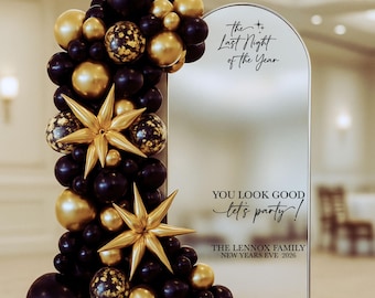 May include: An arched mirror sign with black text reading The Last Night of the Year, YOU LOOK GOOD let's party!, and THE LENNOX FAMILY NEW YEARS EVE 2026, decorated with a dense balloon garland in matte black, metallic gold, and gold confetti.