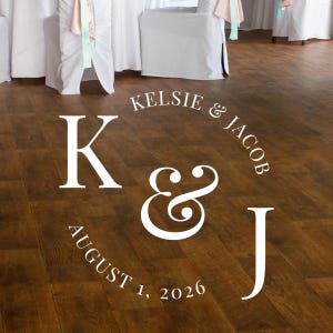 May include: White lettering on a dark wood floor announces a wedding. The text includes the names Kelsie & Jacob, the date August 1, 2026, and the initials K & J. Wedding decor.