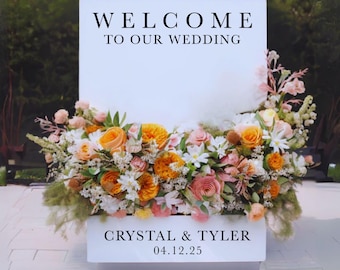 May include: A white rectangular wedding welcome sign featuring the black text "WELCOME TO OUR WEDDING" at the top and "CRYSTAL & TYLER 04.12.25" at the bottom, adorned with a dense arrangement of orange, peach, and white flowers and greenery.