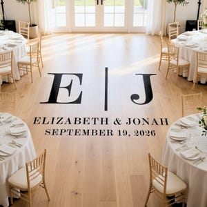May include: A light wood floor featuring a large black vinyl decal with the letters E and J separated by a vertical line, followed by the text ELIZABETH & JONAH and the date SEPTEMBER 19, 2026, surrounded by round tables with white linens.