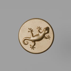 May include: A circular gold-toned medallion featuring a raised, embossed lizard figure in the center. The surface of the disc has a smooth, matte texture, while the lizard has a slightly polished, metallic finish with visible limbs and a curled tail.