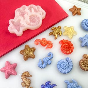 May include: A collection of colorful, miniature sea creature and shell molds and castings. The image includes a starfish, seahorse, turtle, and seashell designs in various colors, including gold, orange, pink, and blue. A silicone mold with similar designs is also present.