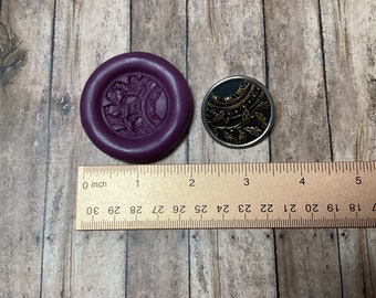 May include: Two round, decorative buttons with a floral design. The button on the left is purple and the button on the right is black with gold accents. A ruler is placed below the buttons to show their size.