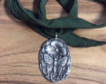 May include: A silver pendant necklace featuring a detailed embossed design of a butterfly and flowers. The necklace is displayed on a green ribbon.