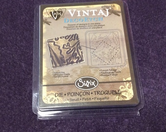 May include: A close-up of a packaging for a die-cutting template for crafting. The template is for a forest foliage design. The packaging is brown and gold with the text "VINTAJ DECOFTCHI" and "Sizzix".