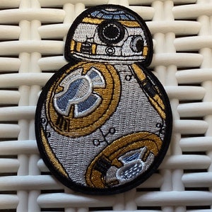 May include: A white, gold, and silver embroidered patch of a Star Wars BB-8 droid. The patch is circular and has a black outline.