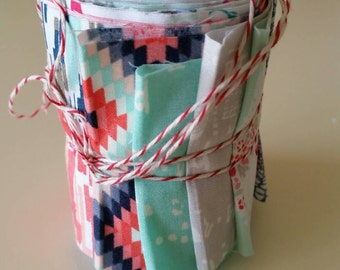 May include: A cylindrical roll of assorted fabric strips featuring geometric and floral patterns in shades of mint green, coral pink, navy blue, and white. The fabric bundle is secured with a thin red and white baker's twine tied in a bow.