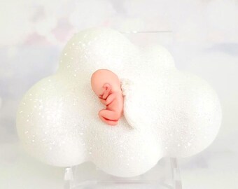 Könnte beinhalten: Ein weißes, wolkenförmiges Ornament mit Glitzerakzenten, das eine kleine, rosa Fötusfigur hält. Die Figur ist zusammengerollt, was ein Baby andeutet. Das Ornament steht auf einem transparenten Ständer vor einem weichen, verschwommenen Hintergrund.
