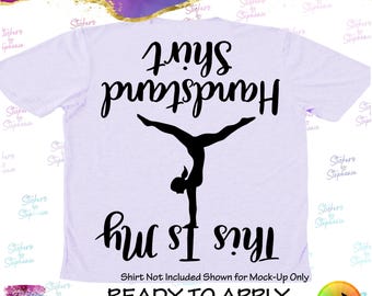 May include: A light purple t-shirt featuring a black silhouette of a person performing a handstand. The text "Handstand Shirt" is printed above the figure, and "This Is My" is printed below, both in a black script font and oriented upside down.