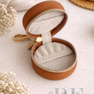 May include: A round, open, brown jewelry case with a gold zipper and clasp. The interior is lined with light gray fabric. The case is on a light-colored surface, with white flowers and the text "Refined by Elegance" in the background.
