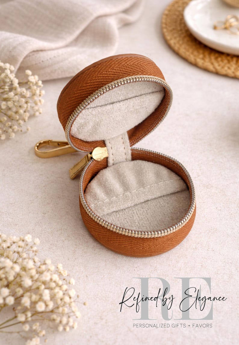 May include: A round, open, brown jewelry case with a gold zipper and clasp. The interior is lined with light gray fabric. The case is on a light-colored surface, with white flowers and the text "Refined by Elegance" in the background.