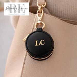 May include: A black round keychain with gold zipper and hardware, featuring the gold initials "LC". The keychain is attached to a beige handbag. The text "Refined by Elegance" is in the upper left corner.