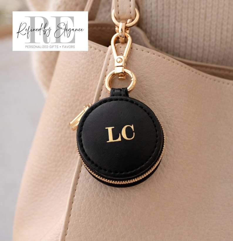 May include: A black round keychain with gold zipper and hardware, featuring the gold initials "LC". The keychain is attached to a beige handbag. The text "Refined by Elegance" is in the upper left corner.