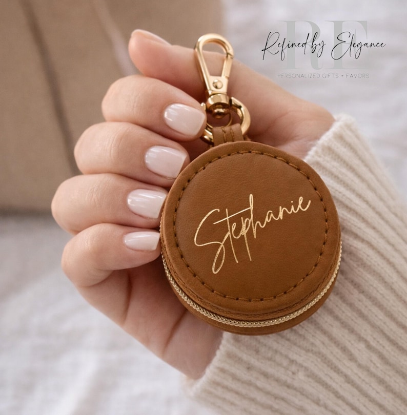 May include: A brown leather round keychain with a gold zipper and clasp. The name "Stephanie" is written in gold script on the front. The keychain is held by a person wearing a beige sweater. The text "Refined by Elegance" is in the background.