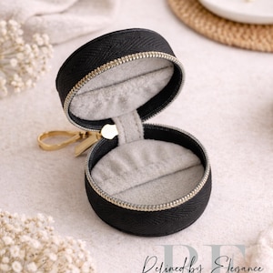 May include: Black round jewelry case with a gold zipper and pull. The interior is lined with gray fabric. The case is open, revealing the inside. The text "Refined by Elegance" is visible in the image.