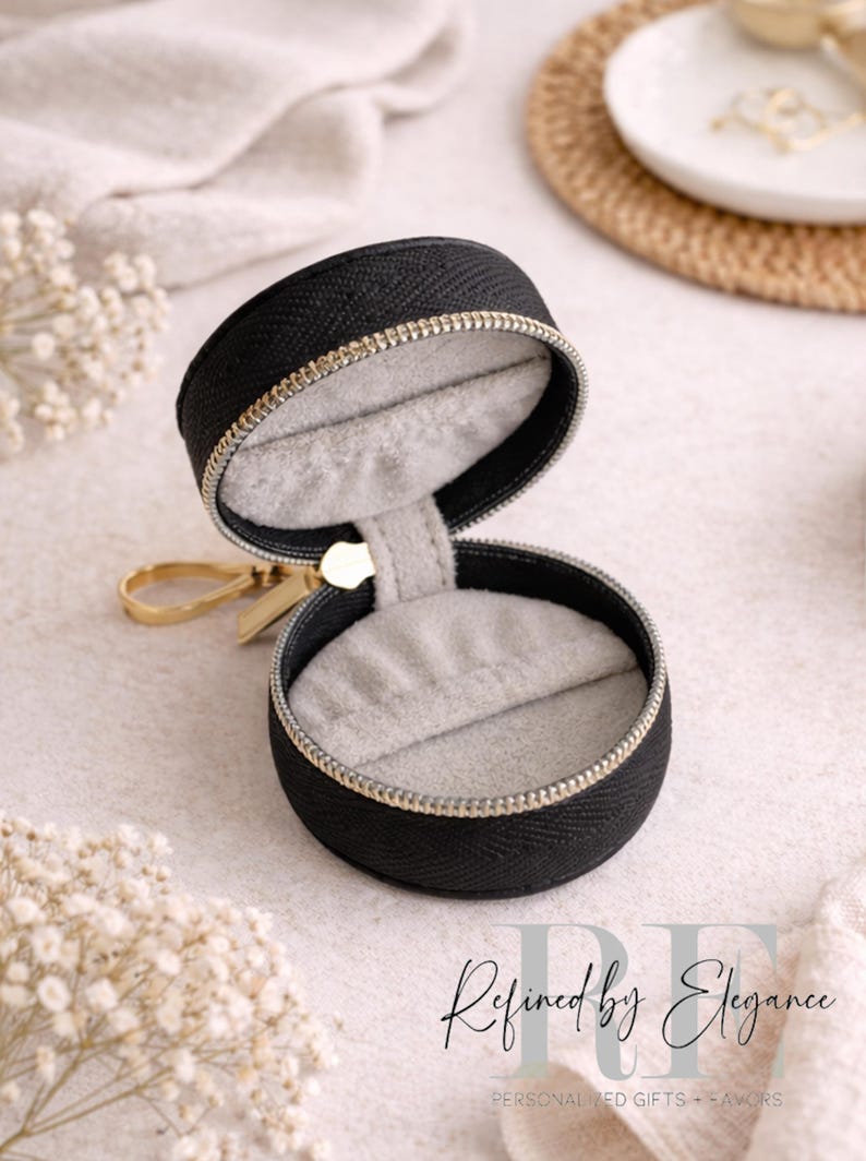 May include: Black round jewelry case with a gold zipper and pull. The interior is lined with gray fabric. The case is open, revealing the inside. The text "Refined by Elegance" is visible in the image.