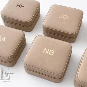 May include: Several beige square jewelry boxes with gold initials are displayed. Each box has a zipper closure and a smooth, leather-like texture. The initials "BP", "DA", "JG", and "NB" are visible.