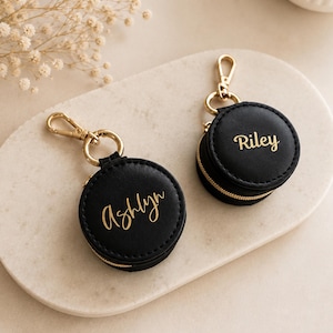 May include: Two round black zippered cases with gold hardware and gold lobster claw clasps. Each case features a name embossed in gold script lettering, with one case reading Ashlyn and the other case reading Riley on the front surface.