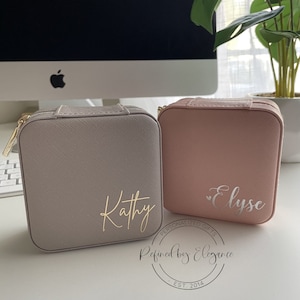 May include: Two personalized jewelry boxes, one in a light gray color with the name "Kathy" in gold lettering and the other in a light pink color with the name "Elyse" in silver lettering. Both boxes have a zipper closure and a textured finish. The boxes are sitting on a white surface with a computer monitor in the background. The text "Personalized Gifts" and "Refined by Elegance" is visible on the bottom of the image.