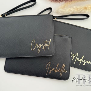 May include: Black clutch purses with gold lettering spelling out names: Crystal, Isabella, and Madison. Each clutch has a wrist strap and a zipper closure. The purses are arranged on a white surface.