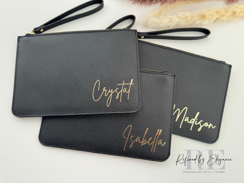 May include: Black clutch purses with gold lettering spelling out names: Crystal, Isabella, and Madison. Each clutch has a wrist strap and a zipper closure. The purses are arranged on a white surface.