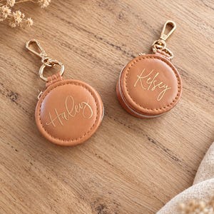 May include: Two round, tan-colored, zippered pouches with gold-toned metal lobster claw clasps. Each pouch features a smooth, leather-like texture and is personalized with gold cursive lettering, one reading Haley and the other reading Kelsey.