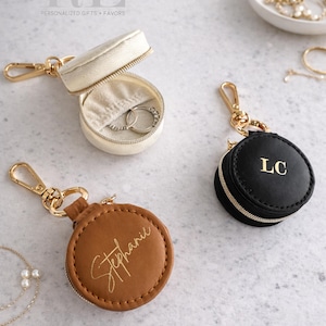 May include: Three small, round, zippered jewelry cases with gold-tone hardware. One is cream-colored and open, revealing rings. Another is brown with "Stephanie" in gold script. The third is black with "LC" in gold. The text "Refined by Elegance" is at the top.