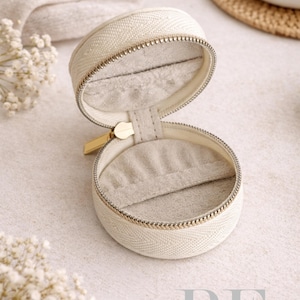 May include: Open, cream-colored, round jewelry case with a gold zipper and a soft, gray interior. The case is made of a textured fabric and sits on a light-colored surface. The text "Refined by Elegance" is visible.