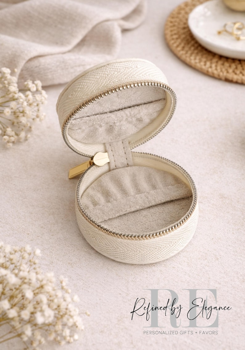 May include: Open, cream-colored, round jewelry case with a gold zipper and a soft, gray interior. The case is made of a textured fabric and sits on a light-colored surface. The text "Refined by Elegance" is visible.