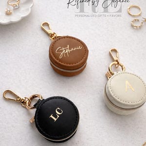 May include: Three personalized round jewelry cases in brown, black, and cream colors. Each case has a gold zipper and a gold-colored clasp. The brown case has the name "Stephanie" in gold lettering. The black case has the initials "LC". The cream case has the letter "A".