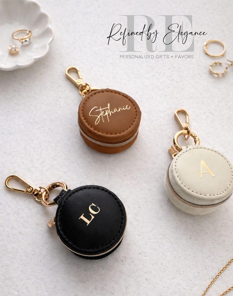 May include: Three personalized round jewelry cases in brown, black, and cream colors. Each case has a gold zipper and a gold-colored clasp. The brown case has the name "Stephanie" in gold lettering. The black case has the initials "LC". The cream case has the letter "A".