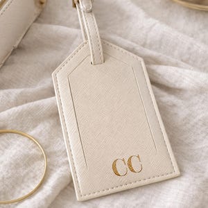 May include: Cream-colored luggage tag with a textured surface and a stitched border. The tag features the gold initials "CC" and a matching strap. A gold-colored bracelet and a small purse are also visible.